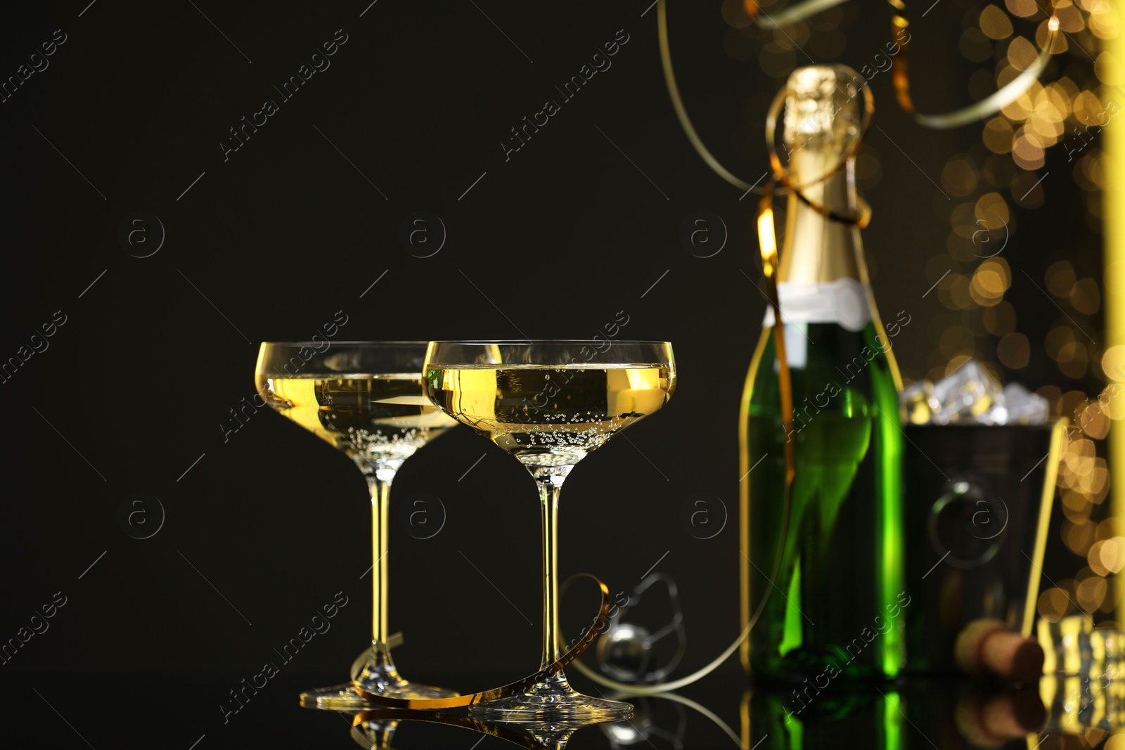 Photo of Champagne in glasses and ice bucket with bottle on mirror surface against black background with blurred lights