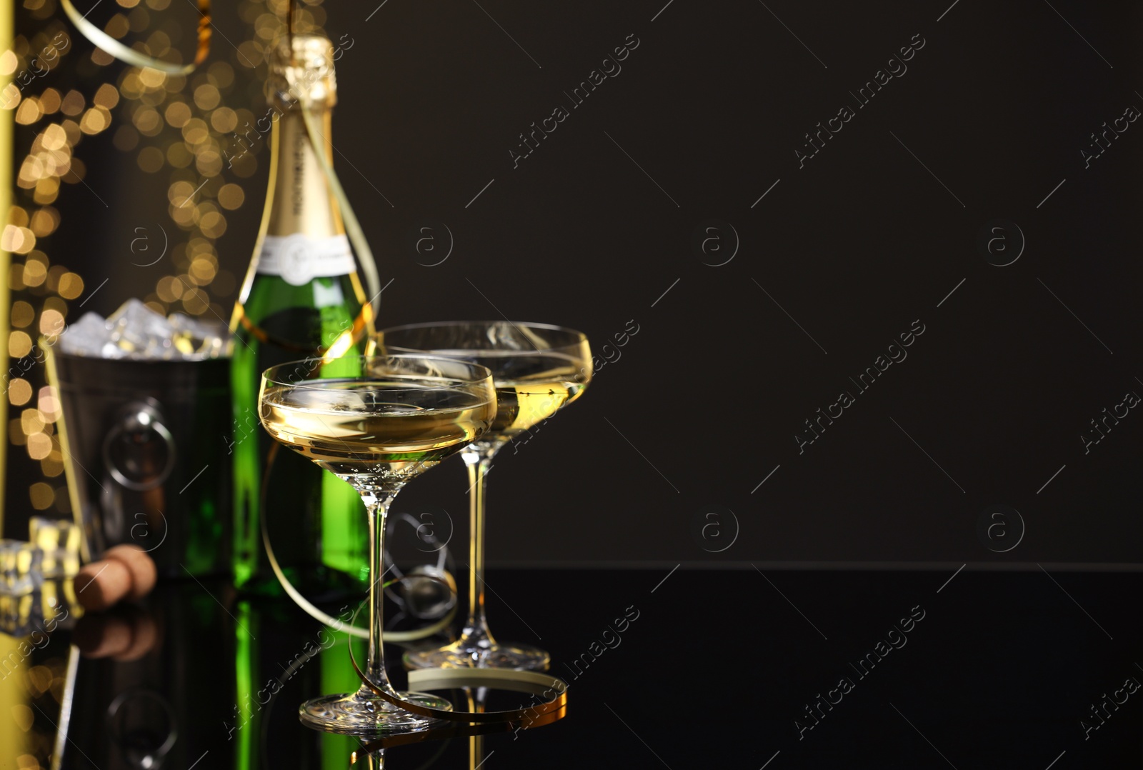 Photo of Champagne in glasses and ice bucket with bottle on mirror surface against black background with blurred lights, space for text