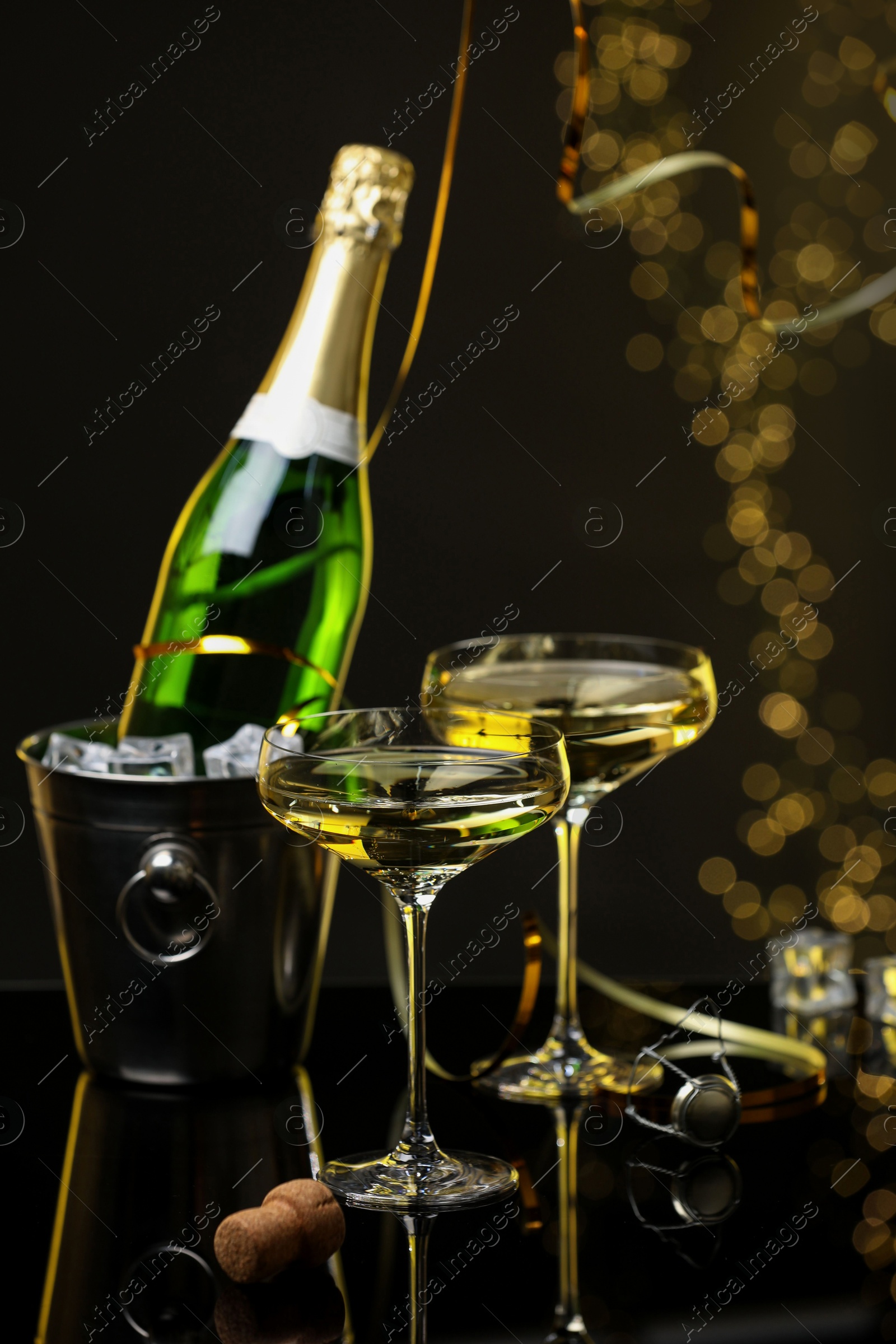 Photo of Champagne in glasses and ice bucket with bottle on mirror surface against black background with blurred lights