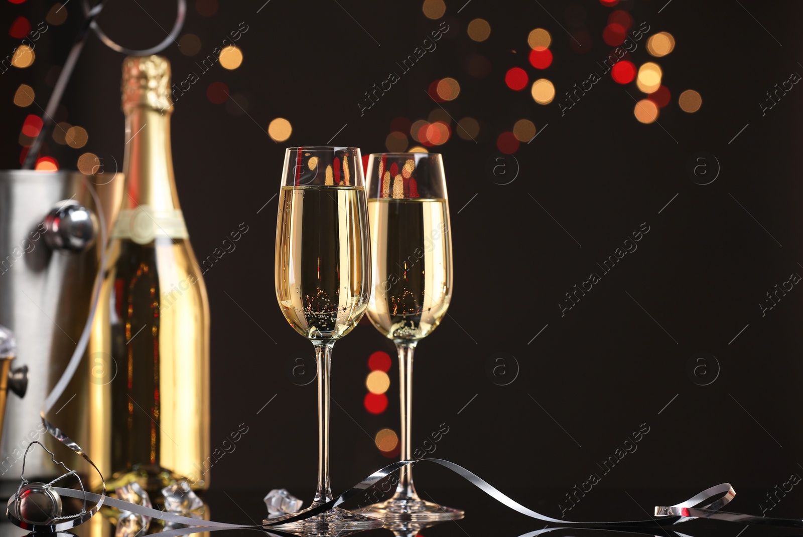 Photo of Champagne in glasses and bottle on mirror surface against black background with blurred lights, space for text