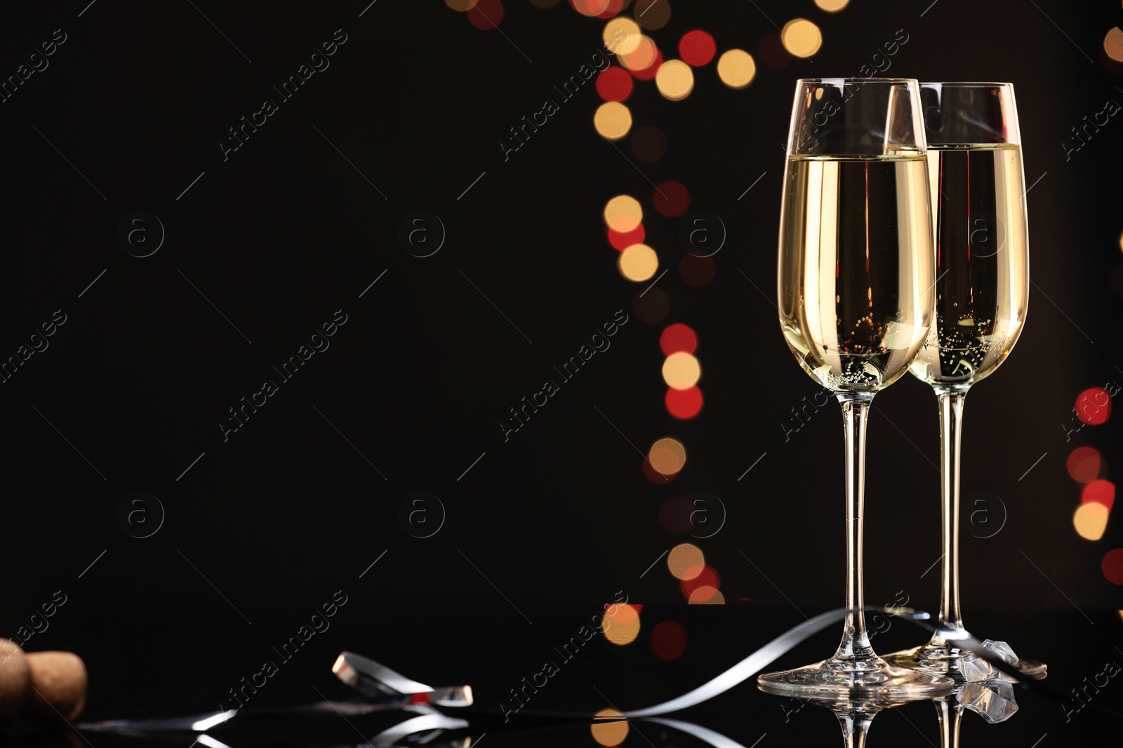 Photo of Champagne in glasses on mirror surface against black background with blurred lights, space for text