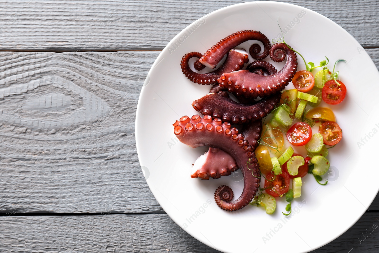 Photo of Plate with tasty boiled octopus tentacles and salad on grey wooden table, top view. Space for text