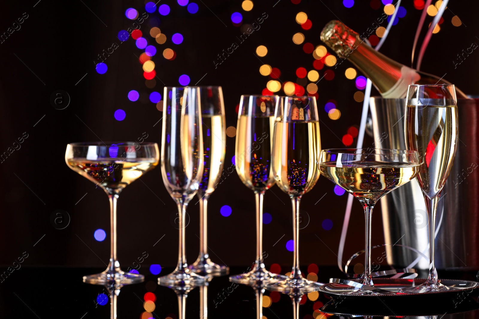 Photo of Champagne in glasses and ice bucket with bottle on mirror surface against dark background with blurred lights