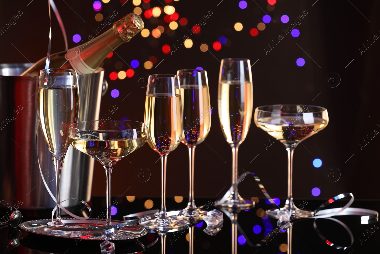 Photo of Champagne in glasses and ice bucket with bottle on mirror surface against dark background with blurred lights