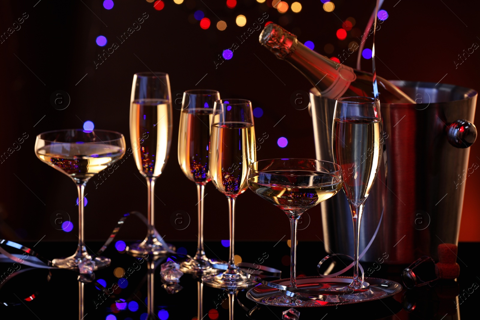 Photo of Champagne in glasses and ice bucket with bottle on mirror surface against dark background with blurred lights