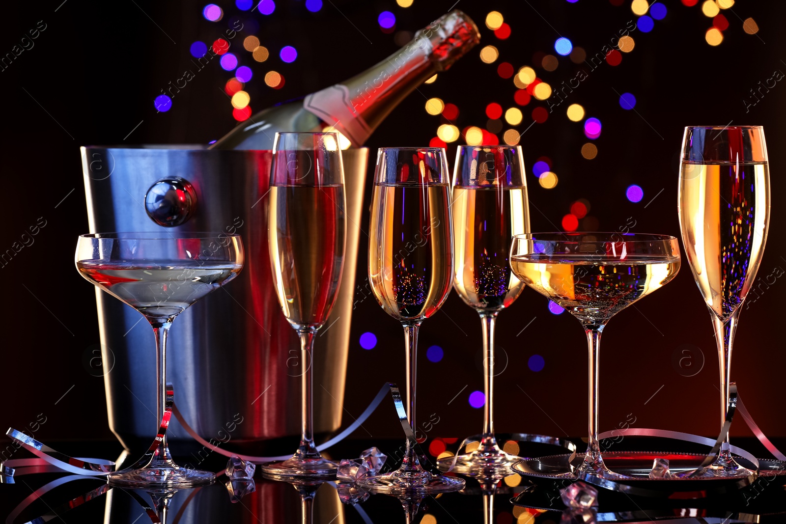 Photo of Champagne in glasses and ice bucket with bottle on mirror surface against dark background with blurred lights