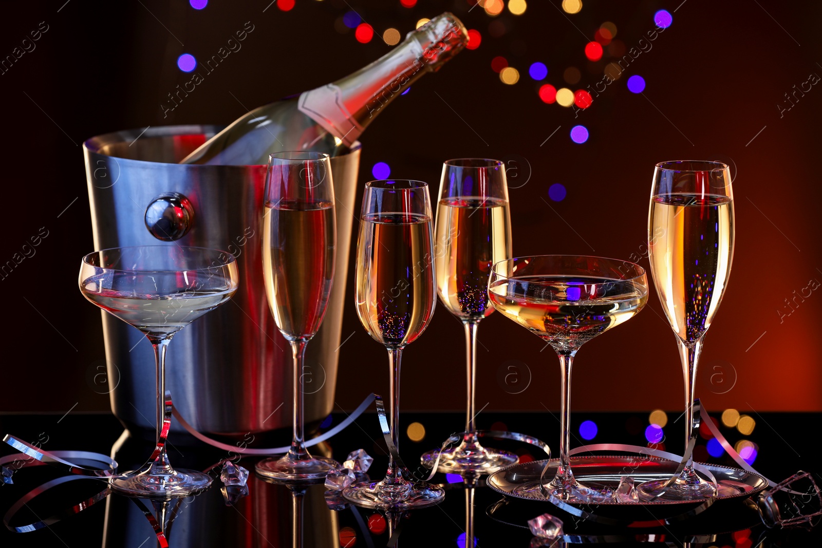 Photo of Champagne in glasses and ice bucket with bottle on mirror surface against dark background with blurred lights