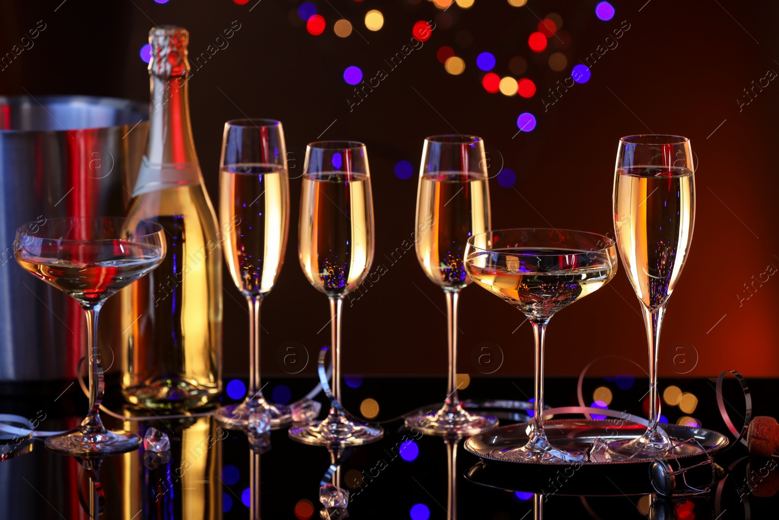 Photo of Champagne in glasses, bottle and ice bucket on mirror surface against dark background with blurred lights