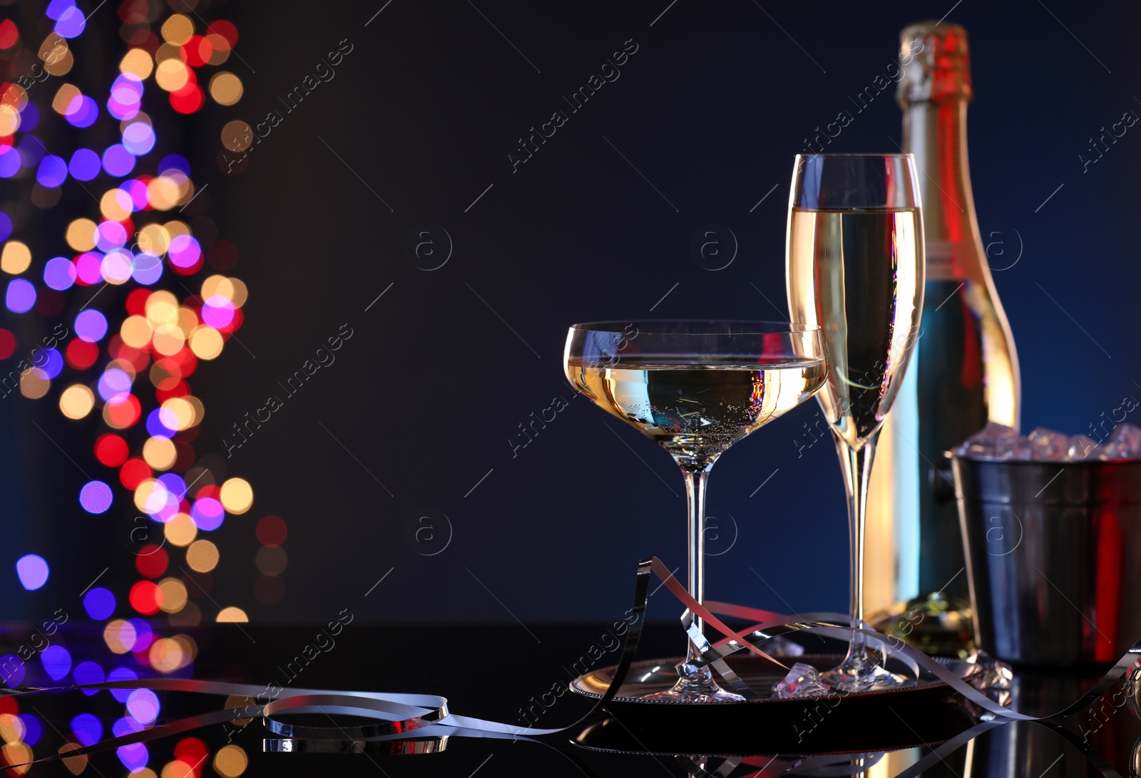 Photo of Champagne in glasses, bottle and ice bucket on mirror surface against dark background with blurred lights, space for text