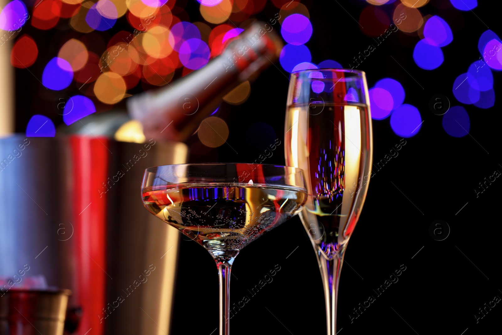 Photo of Champagne in glasses and ice bucket with bottle against black background with blurred lights, closeup