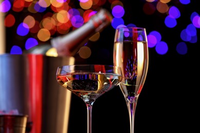 Photo of Champagne in glasses and ice bucket with bottle against black background with blurred lights, closeup