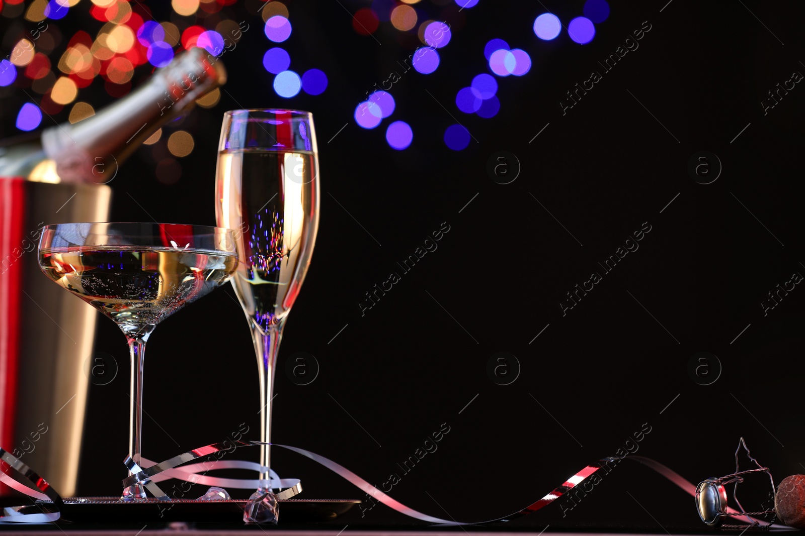 Photo of Champagne in glasses and ice bucket with bottle against black background with blurred lights, space for text