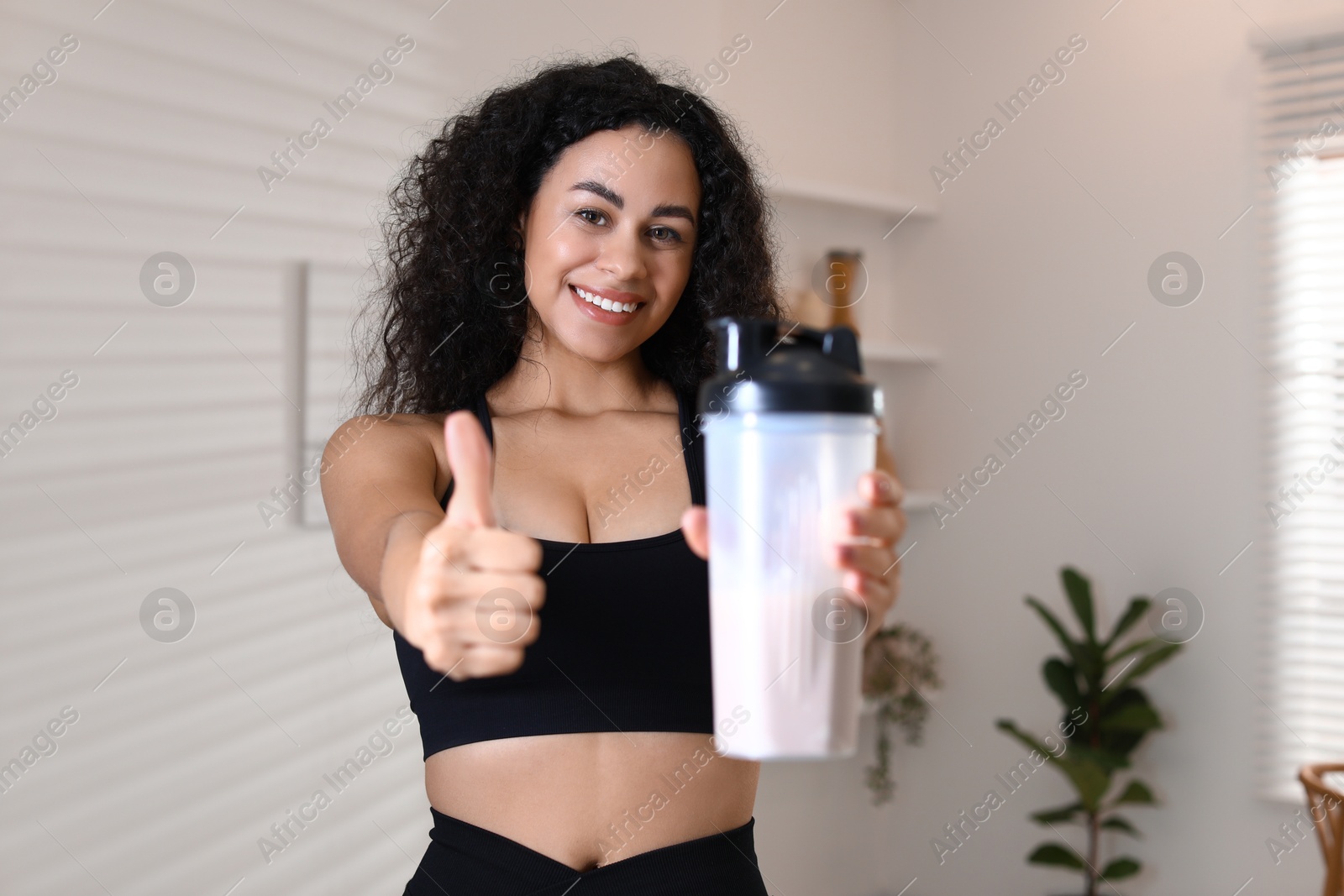 Photo of Beautiful woman with protein shake showing thumbs up at home, selective focus