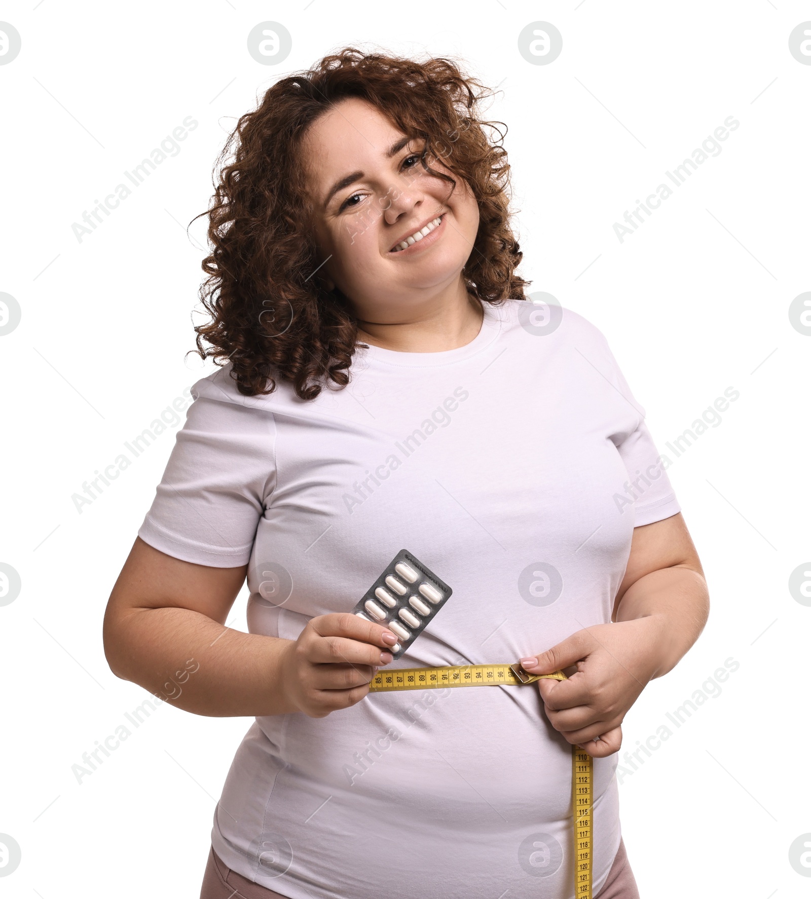 Photo of Happy plus size woman measuring waist with tape and holding blister of weight loss supplements on white background