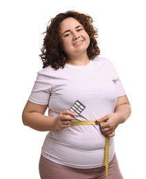 Photo of Happy plus size woman measuring waist with tape and holding blister of weight loss supplements on white background