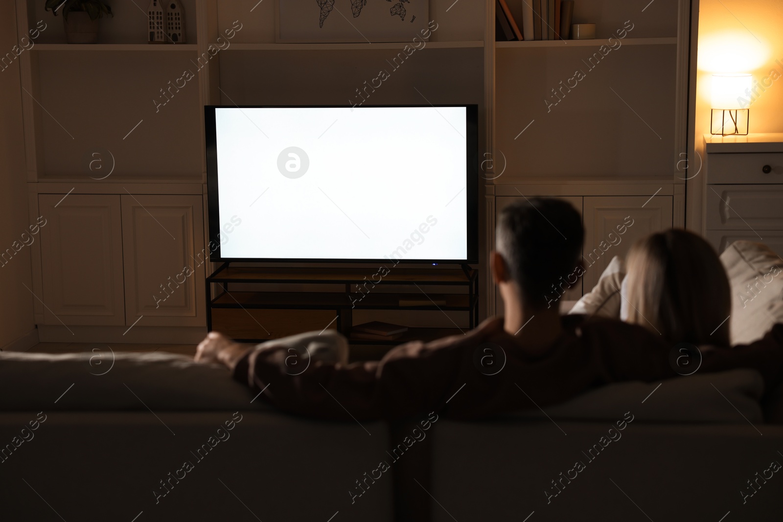 Photo of Couple watching TV on sofa at home in evening, back view