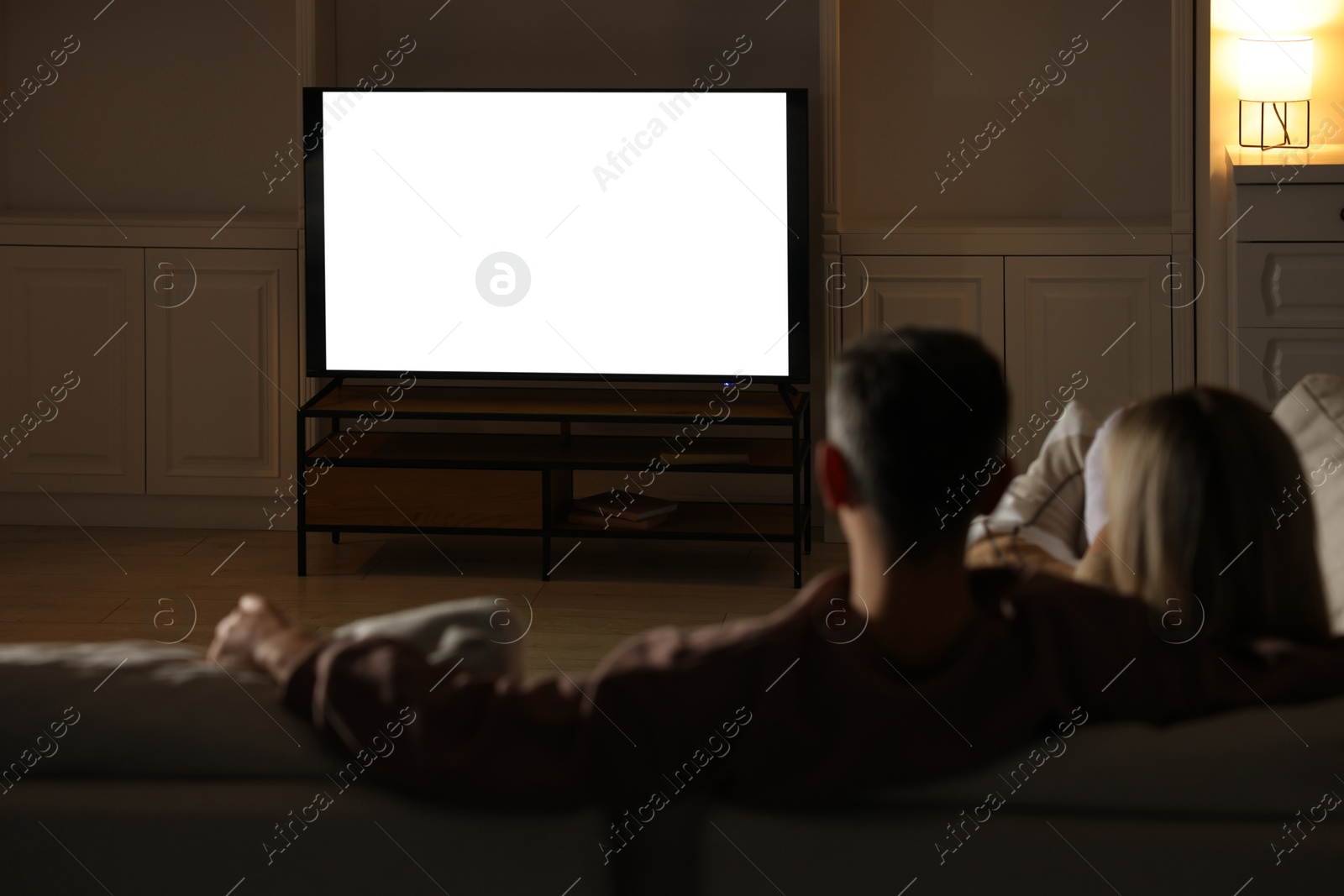 Photo of Couple watching TV on sofa at home in evening, back view