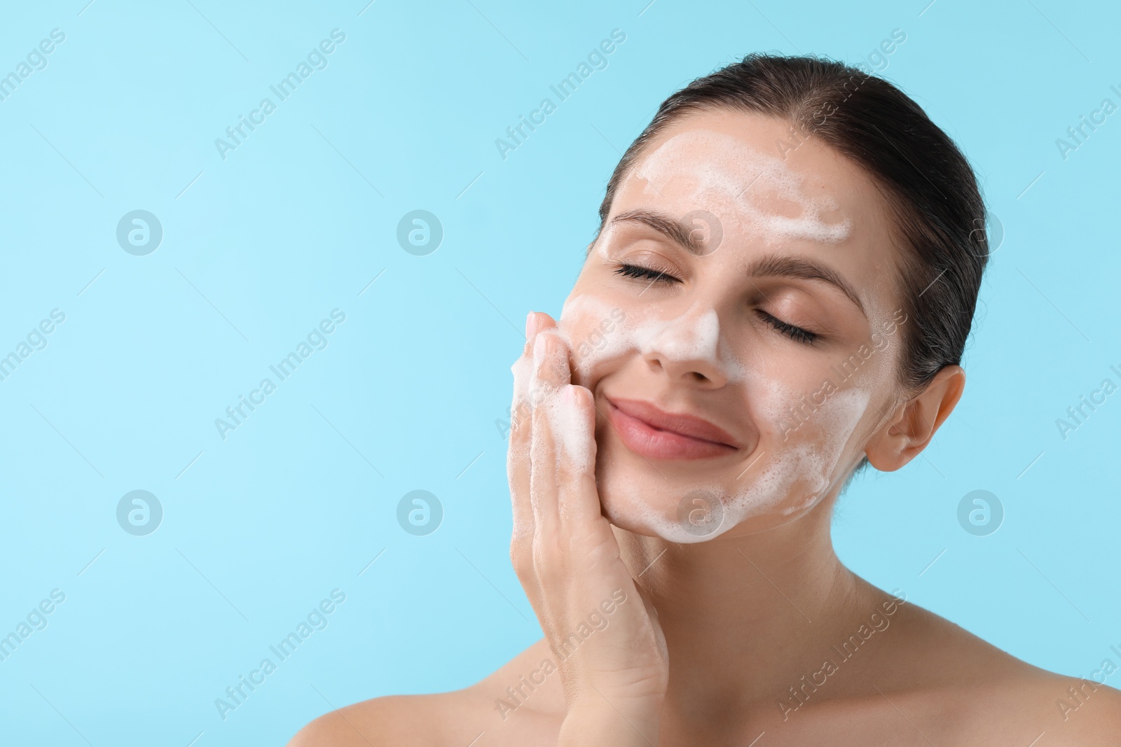 Photo of Attractive woman washing her face with cleansing foam on light blue background, closeup. Space for text