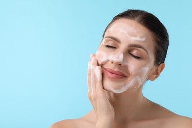 Attractive woman washing her face with cleansing foam on light blue background, closeup. Space for text