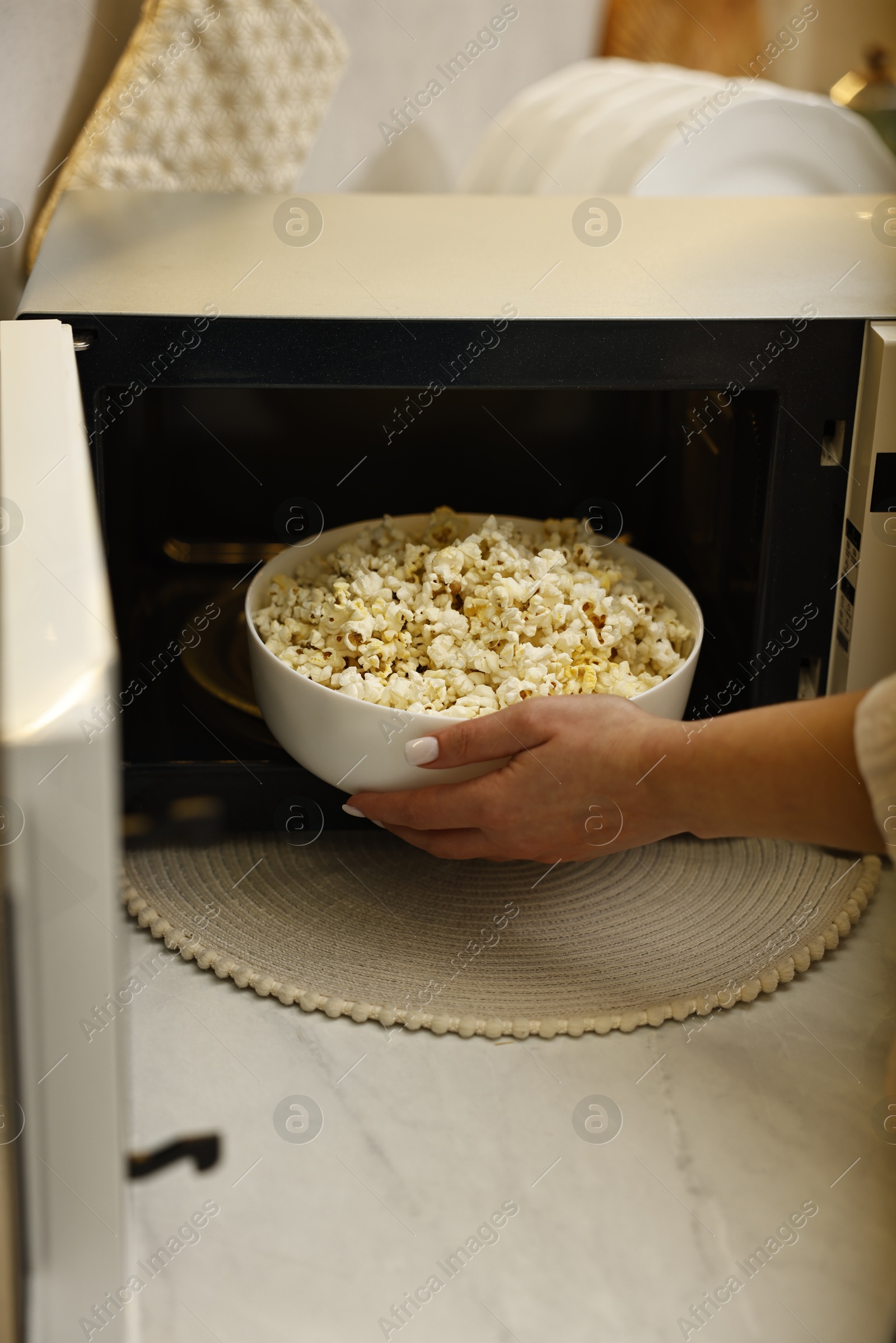 Photo of Woman taking bowl with popcorn out of microwave oven at white marble countertop in kitchen, closeup