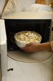 Photo of Woman taking bowl with popcorn out of microwave oven at white marble countertop in kitchen, closeup