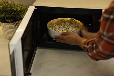 Photo of Woman taking bowl with popcorn out of microwave oven at white marble countertop in kitchen, closeup