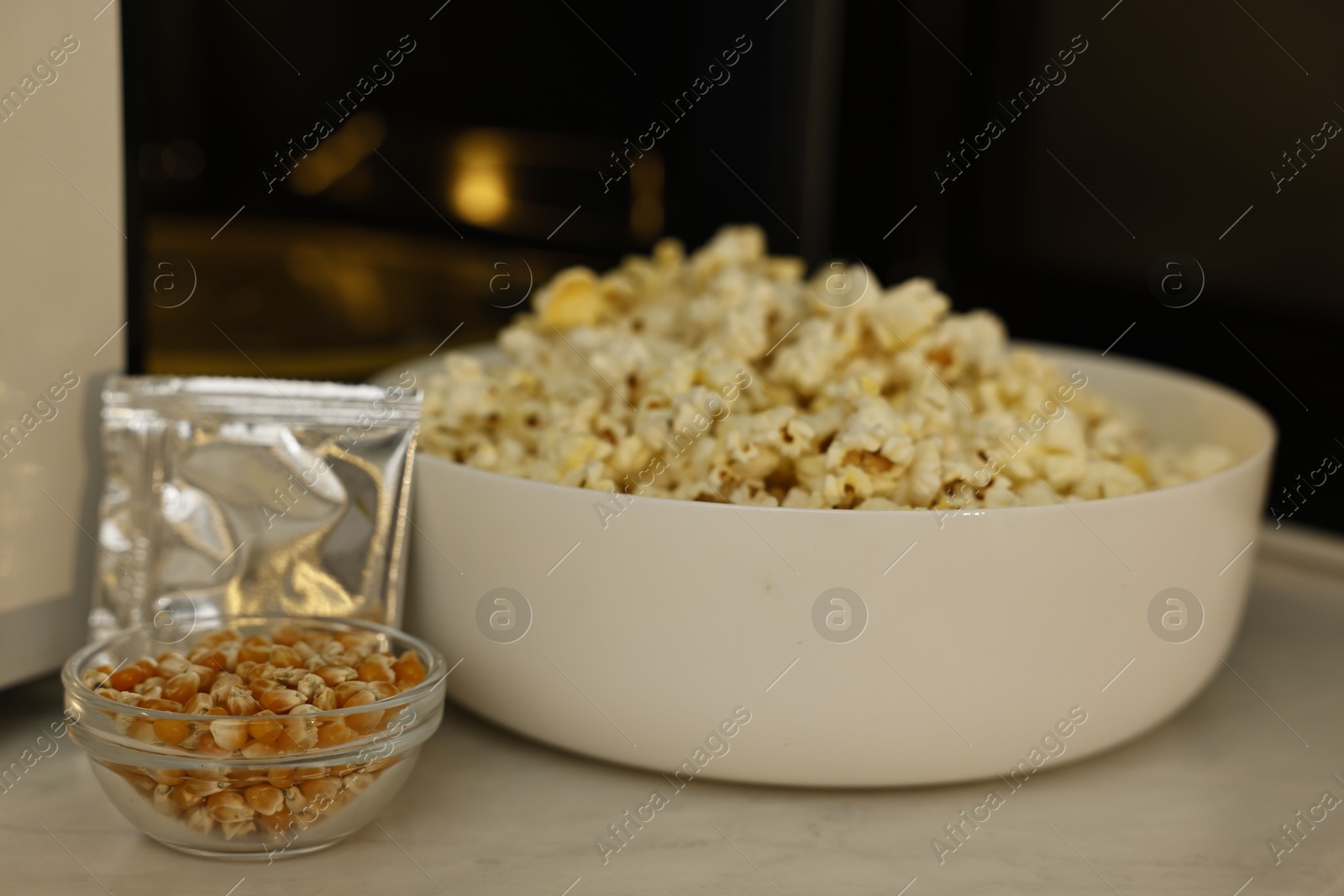 Photo of Bowl of popcorn, kernels and bag near open microwave oven on white marble table, closeup