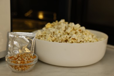 Photo of Bowl of popcorn, kernels and bag near open microwave oven on white marble table, closeup