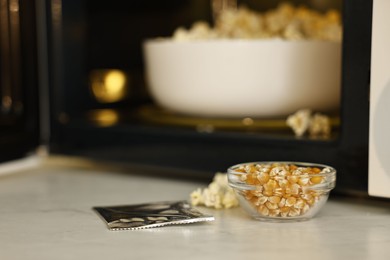 Photo of Unpopped corn kernels in bowl and bag near open microwave oven on white table, closeup. Space for text