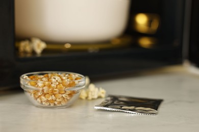 Photo of Unpopped corn kernels in bowl and bag near open microwave oven on white table, closeup. Space for text