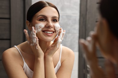 Photo of Woman washing her face with cleansing foam near mirror in bathroom