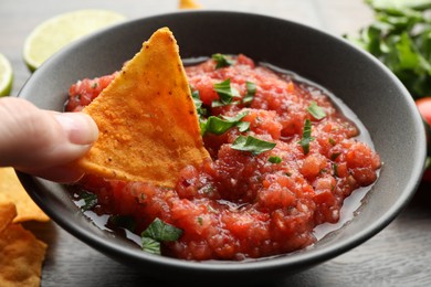 Photo of Woman dipping nacho chip into spicy salsa sauce at wooden table, closeup