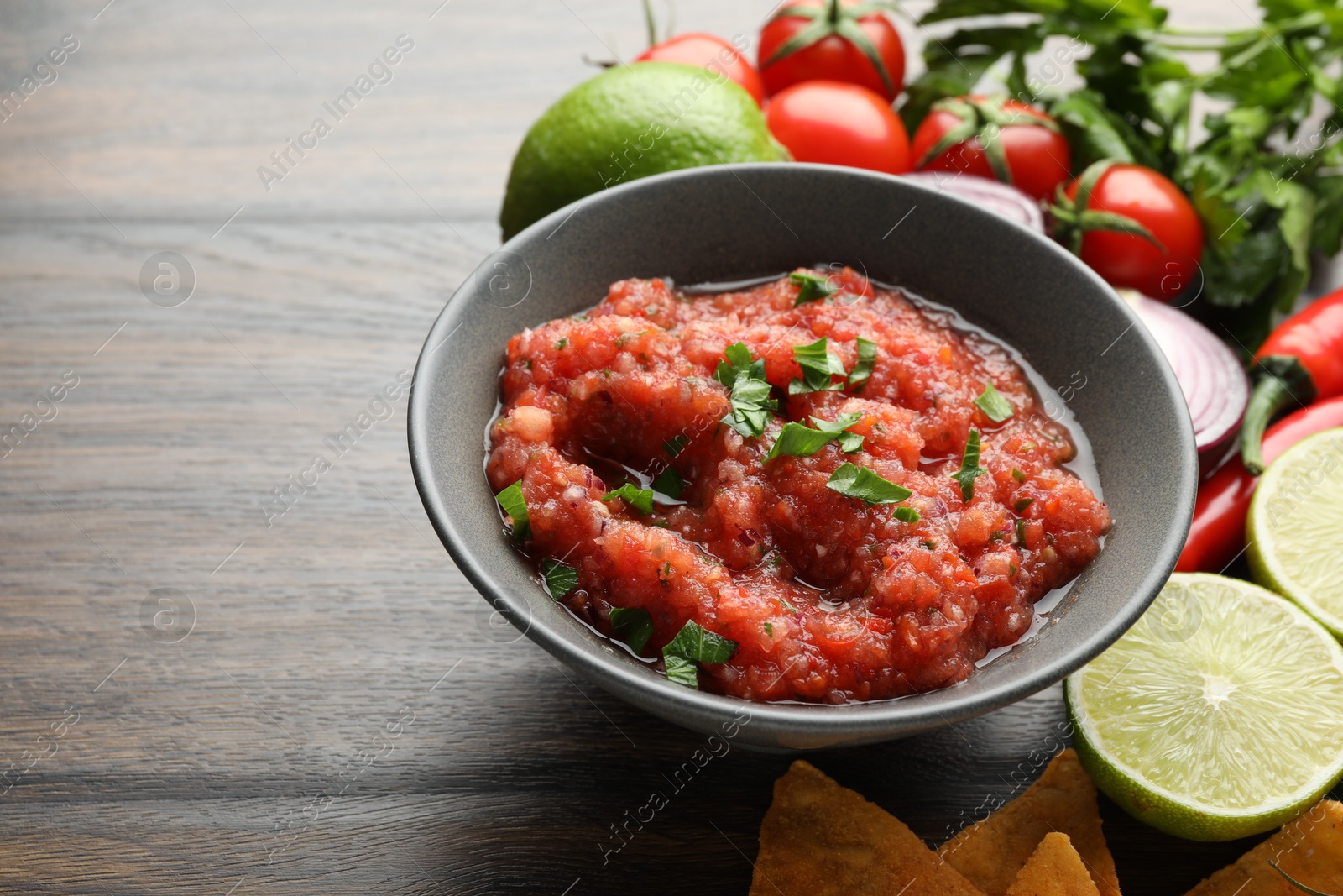 Photo of Delicious spicy salsa sauce in bowl and products on wooden table, closeup. Space for text