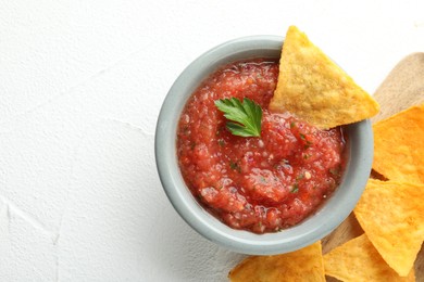 Delicious spicy salsa sauce in bowl and nacho chips on white textured table, flat lay. Space for text