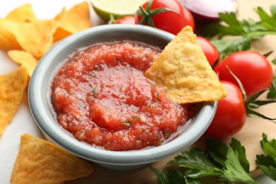 Delicious spicy salsa sauce with nacho chip in bowl and ingredients on white table, closeup