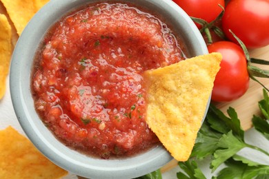 Delicious spicy salsa sauce with nacho chip in bowl and ingredients on white table, flat lay