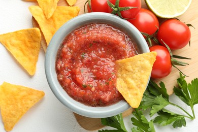 Delicious spicy salsa sauce with nacho chip in bowl and ingredients on white textured table, flat lay