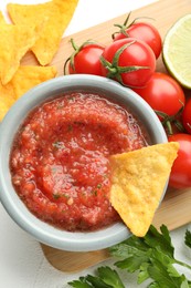 Photo of Delicious spicy salsa sauce with nacho chip in bowl and ingredients on white textured table, flat lay