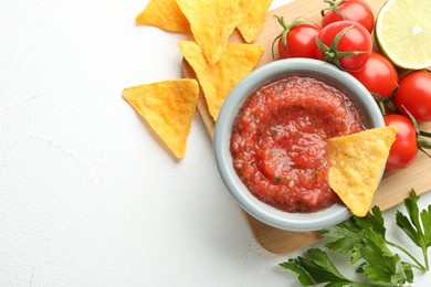 Delicious spicy salsa sauce with nacho chip in bowl and products on white textured table, flat lay. Space for text