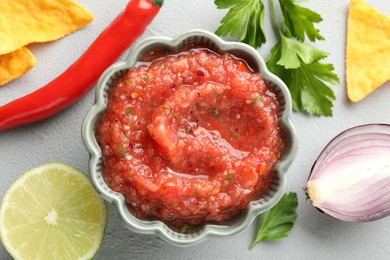 Photo of Delicious spicy salsa sauce in bowl and products on grey textured table, flat lay