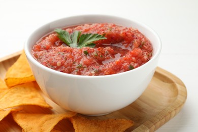 Delicious spicy salsa sauce in bowl and nacho chips on white wooden table, closeup