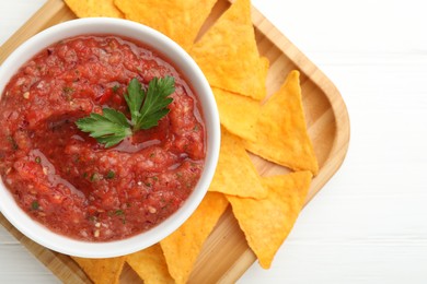 Delicious spicy salsa sauce in bowl and nacho chips on white wooden table, top view