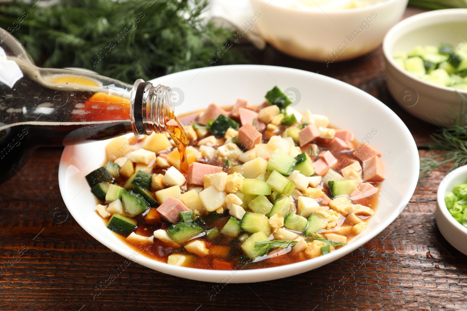 Photo of Making okroshka soup. Pouring kvass into bowl with different ingredients on wooden table, closeup