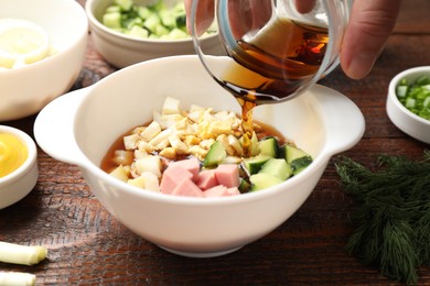 Photo of Making okroshka soup. Woman pouring kvass into bowl with different ingredients at wooden table, closeup