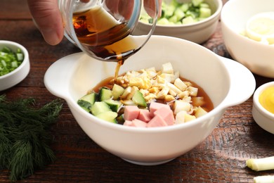 Photo of Making okroshka soup. Woman pouring kvass into bowl with different ingredients at wooden table, closeup