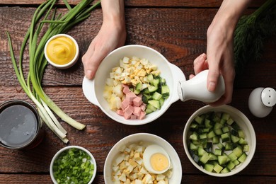 Photo of Making okroshka soup. Woman adding salt into bowl with different ingredients at wooden table, top view