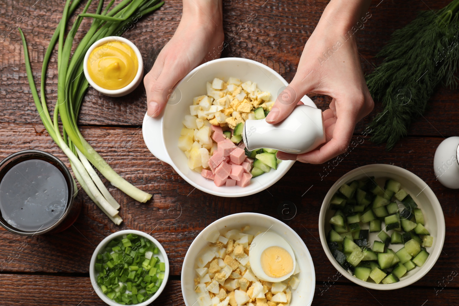 Photo of Making okroshka soup. Woman adding salt into bowl with different ingredients at wooden table, top view