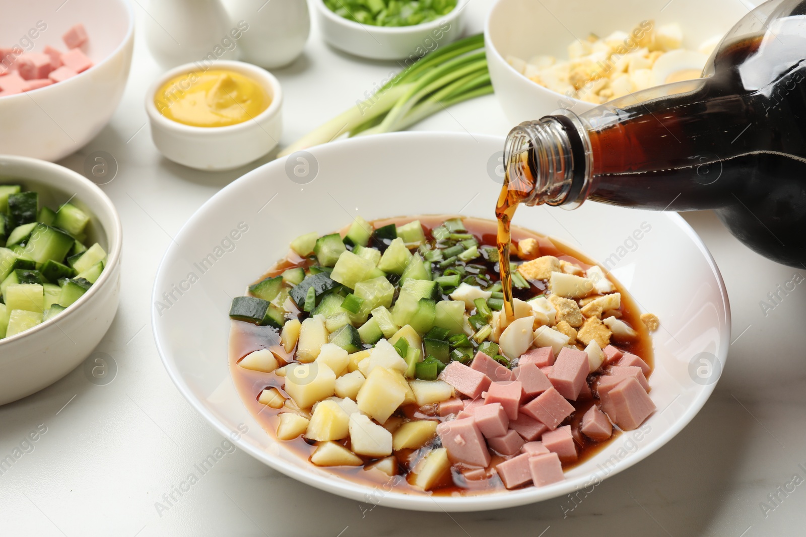 Photo of Making okroshka soup. Pouring kvass into bowl with different ingredients on white marble table, closeup