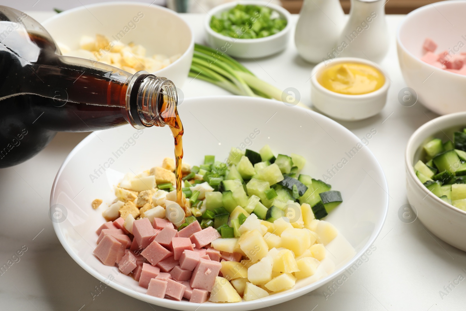 Photo of Making okroshka soup. Pouring kvass into bowl with different ingredients on white marble table, closeup