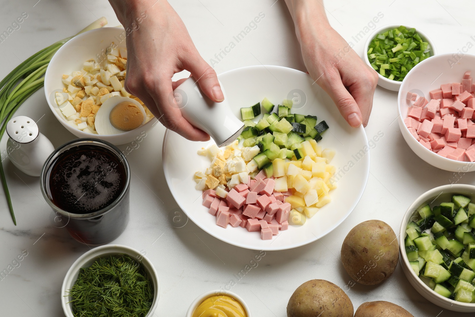 Photo of Making okroshka soup. Woman adding salt into bowl with different ingredients at white marble table, closeup